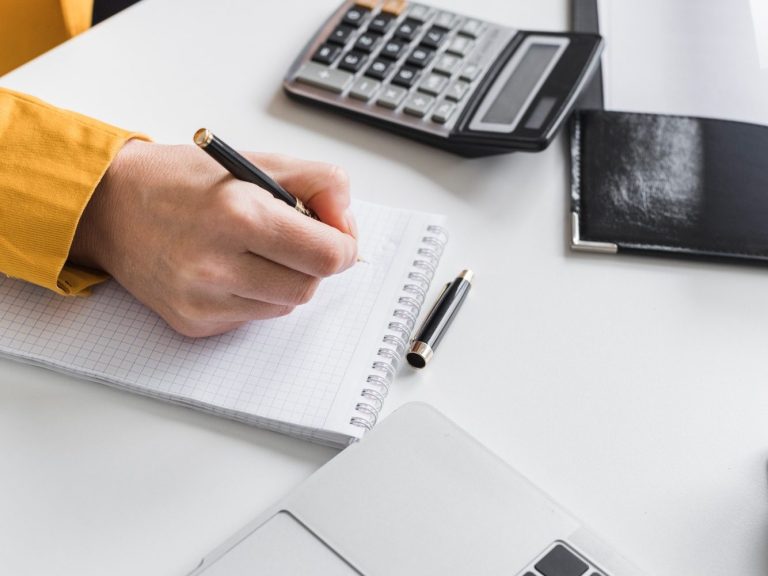 Services aux entrepreneurs Main d'une personne écrivant dans un bloc-notes avec un stylo, calculatrice et documents.